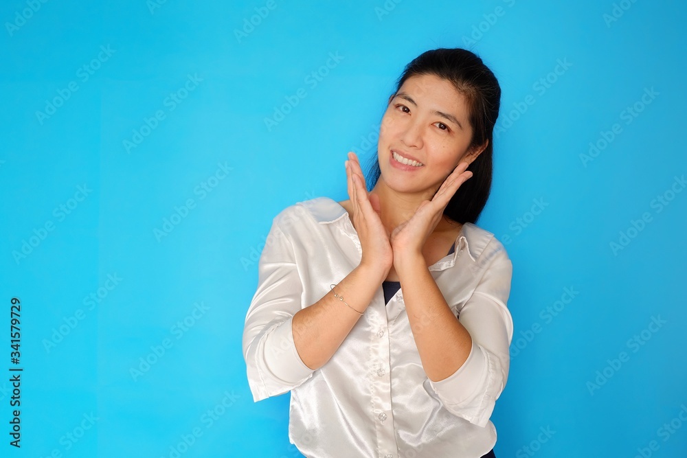 An attractive happy Asian woman posing with her hands by her chin, tilting her head slightly with plain light blue background An attractive happy Asian woman posing with her hands by her chin, tilting her head slightly with plain light blue background