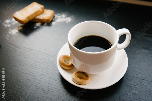 Simple set up for morniing coffee break which includes coffee cup and saucer and few biscuits and bagels plus a bunch of cinnamon sticks. and sugar powder for decoration. black background