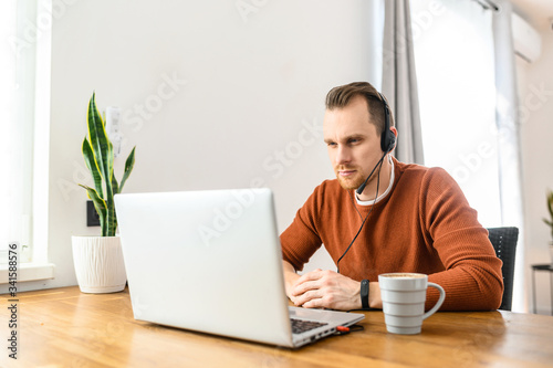 Video conference from home; morning meeting remotely. A guy uses a headset for online communication, he talks using a laptop. Work from home concept