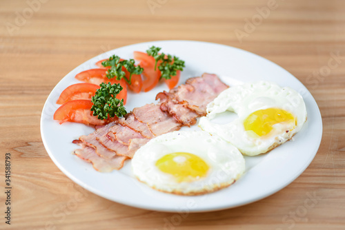 Breakfast with bacon, fried egg and tomatoes on served on white plate over light rustic wooden table.