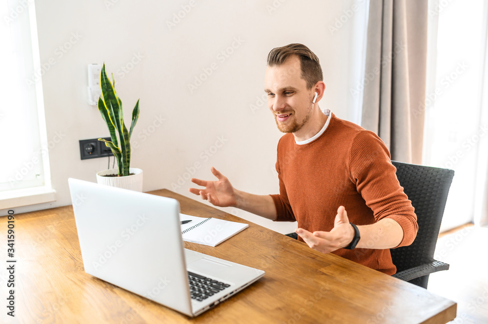 Video call. A young positive guy in casual clothes is using a laptop and airpods for online communication. He sits at the table, looks at the laptop screen and talks. Back view Video call. A young positive guy in casual clothes is using a laptop and airpods for online communication. He sits at the table, looks at the laptop screen and talks. Back view
