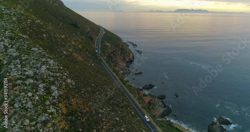 A striking aerial over cars driving along a road at the base of a lush hill by the shoreline of an enormous bay at dusk - Cape Town, South Africa photo