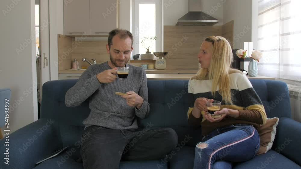 Wife brings coffee to husband, they sit on couch, smile, talk and drink