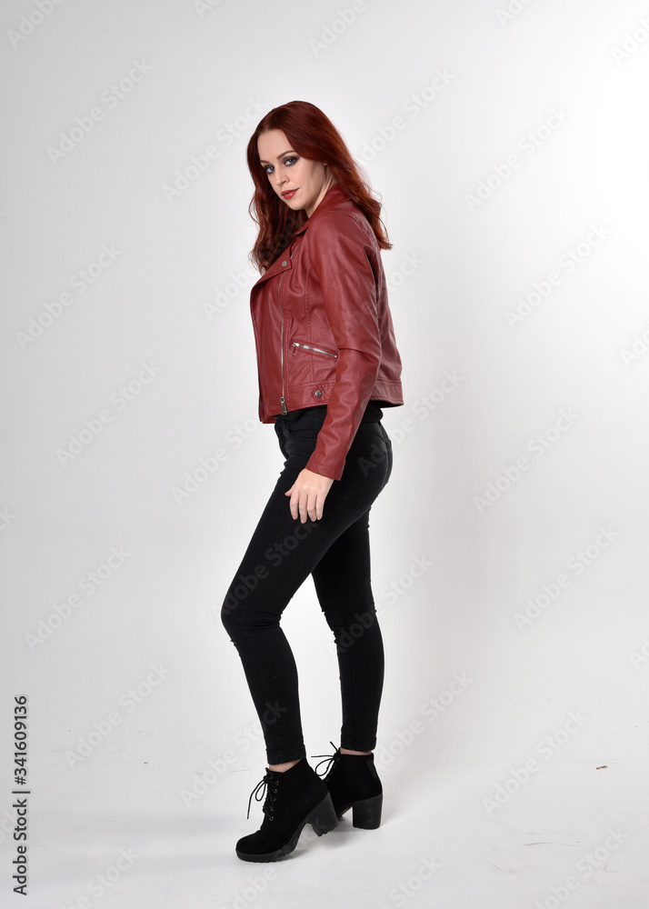 Portrait of a pretty girl with red hair wearing black jeans and boots with leather jacket. full length standing pose with back to the camera a studio background. Portrait of a pretty girl with red hair wearing black jeans and boots with leather jacket. full length standing pose with back to the camera a studio background.
