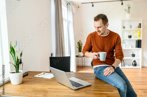 Morning coffee on the workplace. Handsome positive guy using laptop for work, he sits on the edge of table with cup of coffee in hands and looks in screen with a smile