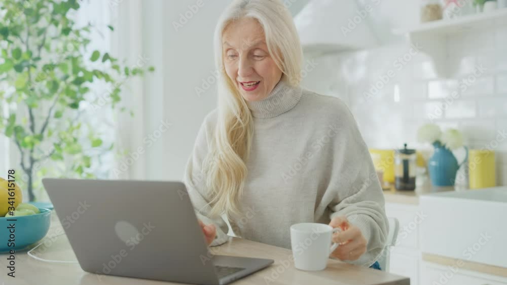 Beautiful Senior Woman Using Laptop Computer and Drinking a Cup of Coffee or Tea in a Sunny Kitchen. Grandmother is Connected with Children via internet and Social Media. Pensioner Working from Home. Beautiful Senior Woman Using Laptop Computer and Drinking a Cup of Coffee or Tea in a Sunny Kitchen. Grandmother is Connected with Children via internet and Social Media. Pensioner Working from Home.