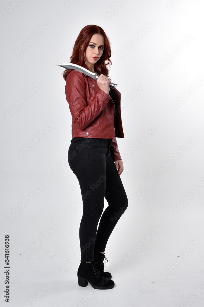 Portrait of a pretty girl with red hair wearing black jeans and boots with leather jacket. full length standing pose holding knife a studio background. Portrait of a pretty girl with red hair wearing black jeans and boots with leather jacket. full length standing pose holding knife a studio background.