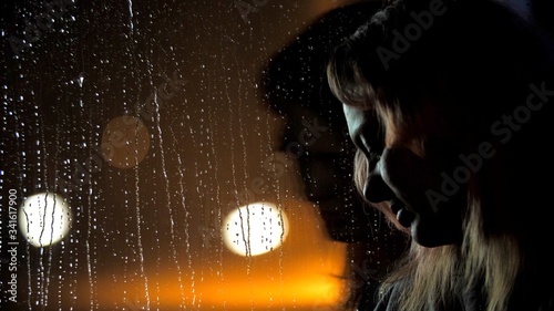 Close up portrait of young attractive sad woman is sitting at night near the window. It is raining outside and the lights of the city. large drops glide across the glass. Self-isolation in quarantine