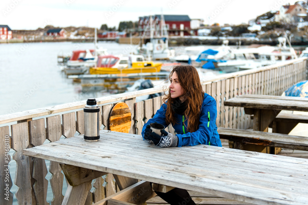 Happy smiling girl traveler drinks warm tea at the rustic wooden table outdoor near the traditional fishing houses Rorbu and fishing ships in Lofoten islands, Norway