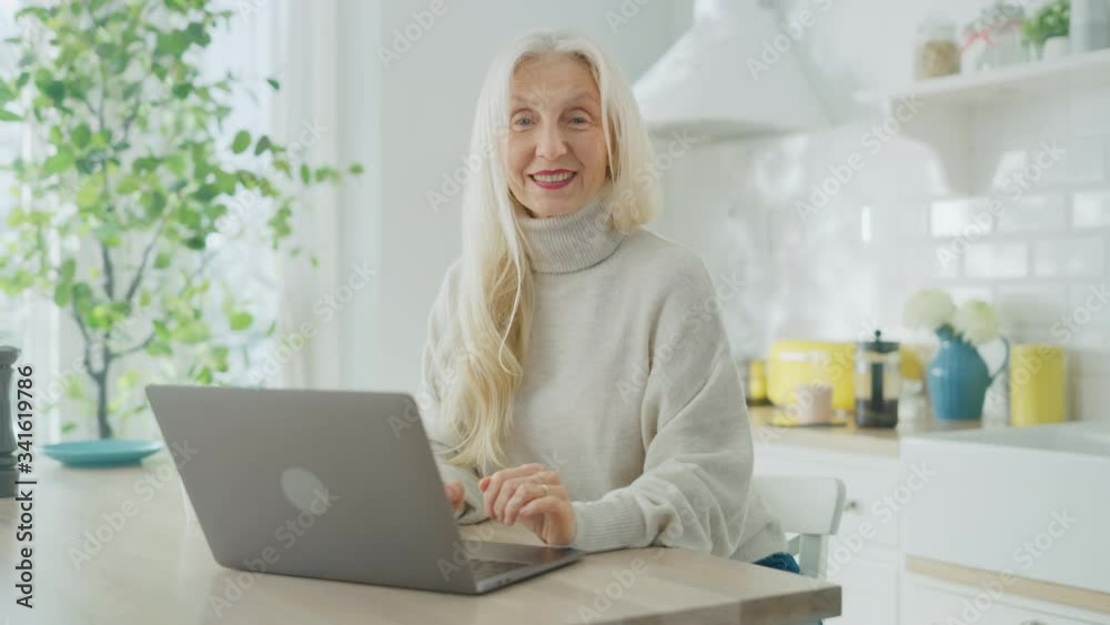 Beautiful Senior Woman Using Laptop Computer in a Sunny Kitchen. She Looks at Camera and Smiles. Grandmother is Connected with Children via internet and Social Media. Pensioner Working from Home. Beautiful Senior Woman Using Laptop Computer in a Sunny Kitchen. She Looks at Camera and Smiles. Grandmother is Connected with Children via internet and Social Media. Pensioner Working from Home.