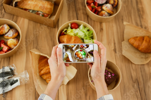Female food blogger reviewing takeout food, taking video on phone. Italian sub sandwich, french croissant w/ salmon, chocolate cheesecake. Close up, top view, pov, copy space, wooden table background.
