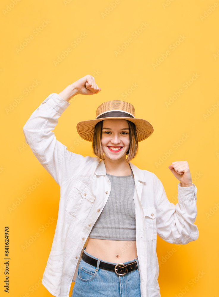 Joyful lady in hat and summer dress rejoices in victory with raised hands and happy face, looks into camera with smile on face, isolated on yellow background. Vertical. Joyful lady in hat and summer dress rejoices in victory with raised hands and happy face, looks into camera with smile on face, isolated on yellow background. Vertical.