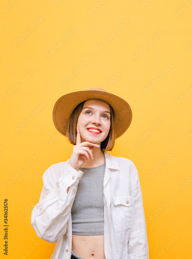 Happy young girl in light summer clothes and hat with thoughtful look looks up at copy space and smiles. Portrait of pensive lady on yellow background, looking up and thinking. Isolated. Happy young girl in light summer clothes and hat with thoughtful look looks up at copy space and smiles. Portrait of pensive lady on yellow background, looking up and thinking. Isolated.