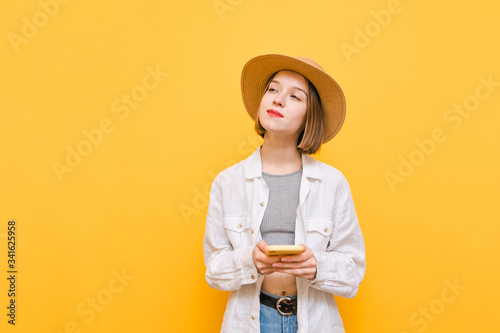 Suspicious pretty girl in summer clothes and hat stands on yellow background with smartphone in hand, looks away with pensive face. Pensive lady isolated. Copy space