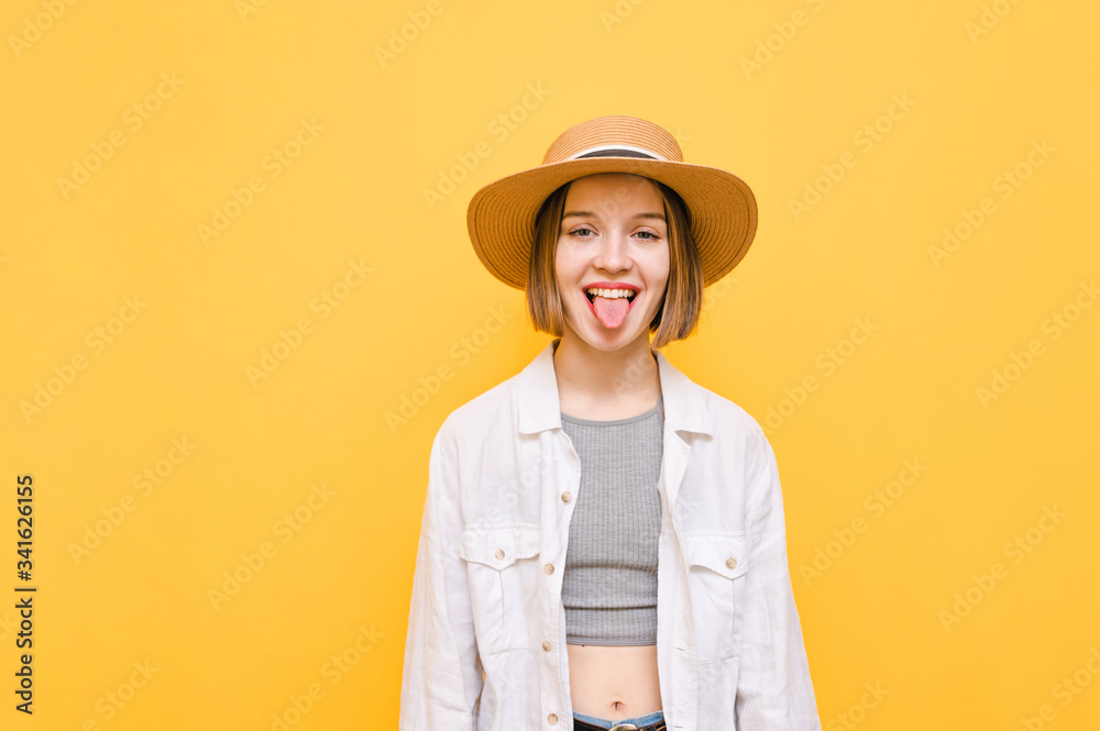 Happy tourist girl in summer clothes and hat isolated on yellow background, looks into camera with smiling face and shows tongue. Funny lady in light clothing shows tongue and laughs, isolated. Happy tourist girl in summer clothes and hat isolated on yellow background, looks into camera with smiling face and shows tongue. Funny lady in light clothing shows tongue and laughs, isolated.