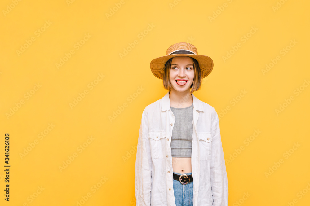 Funny girl in stylish light summer clothes and hat stands on a yellow background, looks to the empty space with a happy face and shows tongue. Isolated. Copy space Funny girl in stylish light summer clothes and hat stands on a yellow background, looks to the empty space with a happy face and shows tongue. Isolated. Copy space