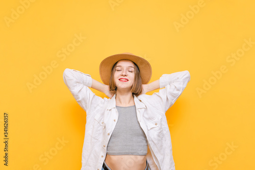 Portrait of a cute smiling girl in a summer dress and sun hat stands on a yellow background with her eyes closed and smiling. Vacationer girl in hat enjoys, isolated on yellow. Copy space