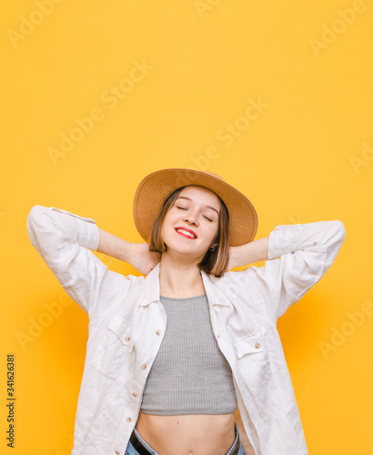 Smiling girl in sun hat and light clothing is resting with eyes closed on yellow background. Attractive lady holds pleasure against yellow background with happy face. Vertical. Copy space