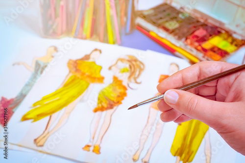 The artist holds a thin brush in his hand against the background of his sketch in watercolors. The picture shows two girls in yellow and orange evening dresses.