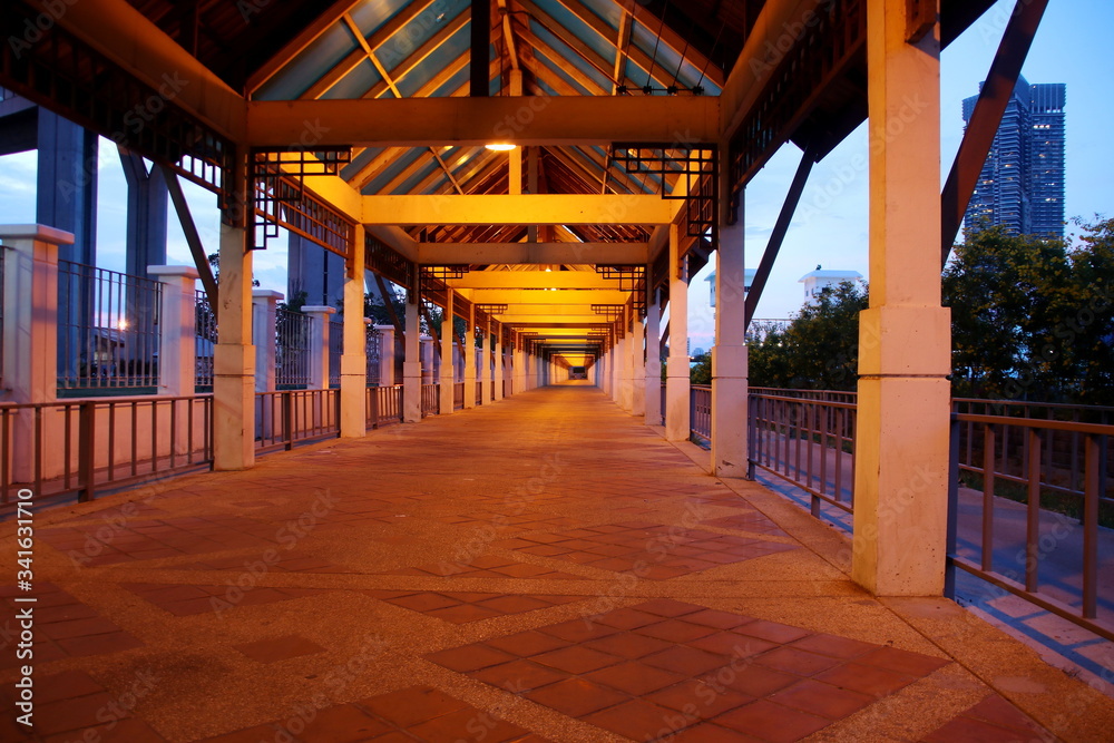 Walkway under triangle roof in the evening with orange light from lamp.  Under roof open structure of building, Thailand.