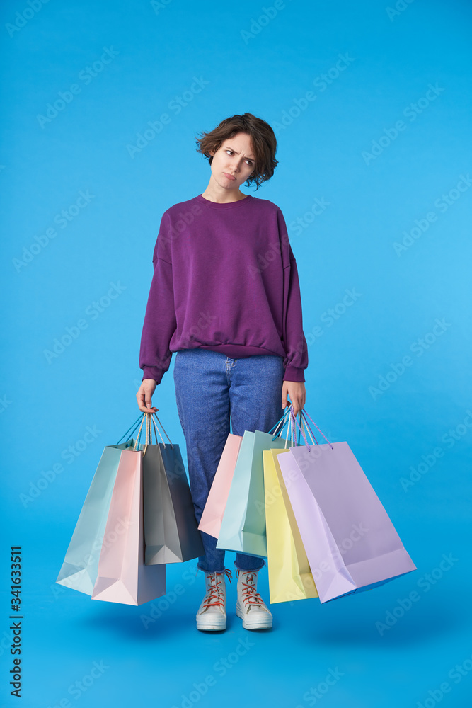 Displeased young dark haired woman with paper bags being tired after shopping and frowning tiredly her face, dressed in casual wear while standing over blue background Displeased young dark haired woman with paper bags being tired after shopping and frowning tiredly her face, dressed in casual wear while standing over blue background