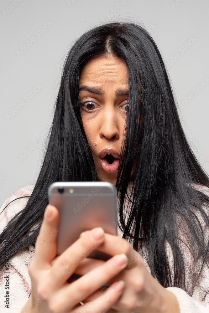 Portrait of an anxious young girl looking at the phone, seeing bad news or photos with disgusting emotions on her face, isolated on a light gray background. Human emotion, reaction, expression.