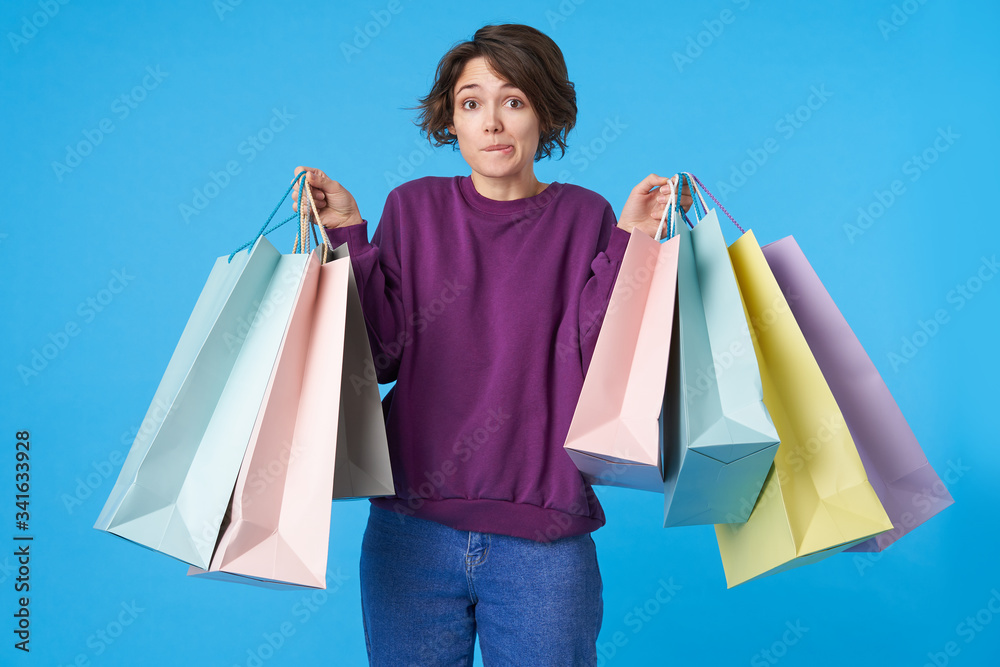 Confused young pretty short haired brunette female with purchases in raised hands looking wonderingly at camera with raised eyebrows, isolated over blue background