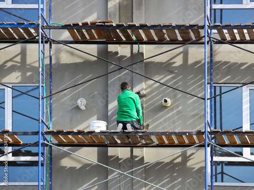 A worker plastering a building facade while standing on a scaffold. Construction work on the construction of a high-rise building. Safety measures for high-rise construction works. Insulation of thin