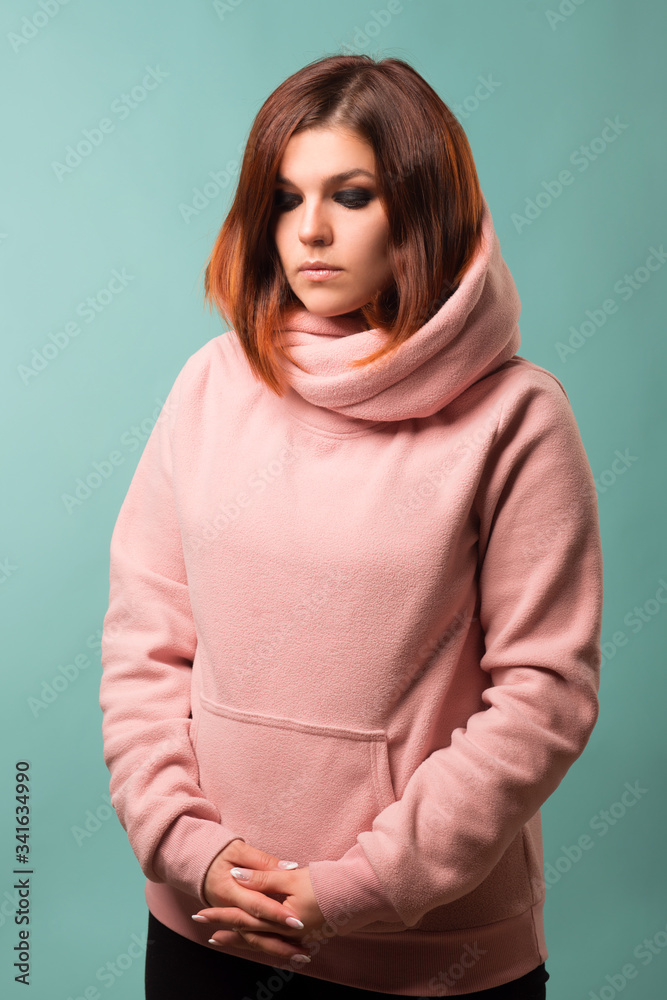 Redhead girl in a pink hoodie looks down on a blue background. Studio woman portrait. Redhead girl in a pink hoodie looks down on a blue background. Studio woman portrait.