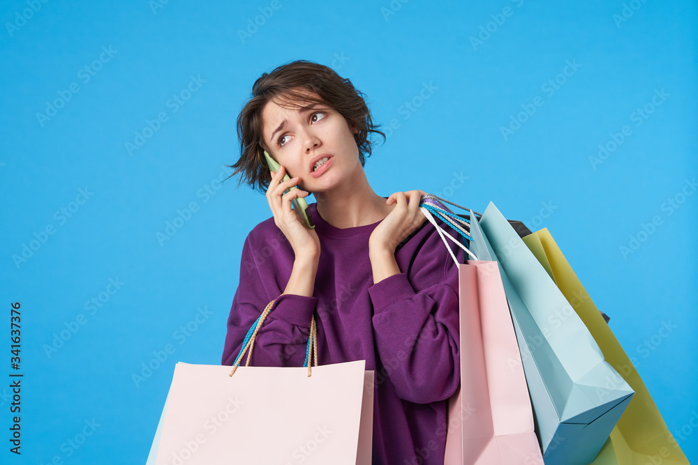Bewildered young short haired pretty female dressed in purple sweatshirt looking confusedly aside while making call, holding shopping bags while standing over blue background Bewildered young short haired pretty female dressed in purple sweatshirt looking confusedly aside while making call, holding shopping bags while standing over blue background