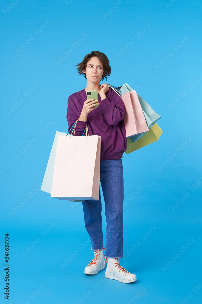 Pensive young pretty curly brunette woman squinting her eyes while looking thoughtfully aside and holding mobile phone, standing over blue background with shopping bags Pensive young pretty curly brunette woman squinting her eyes while looking thoughtfully aside and holding mobile phone, standing over blue background with shopping bags