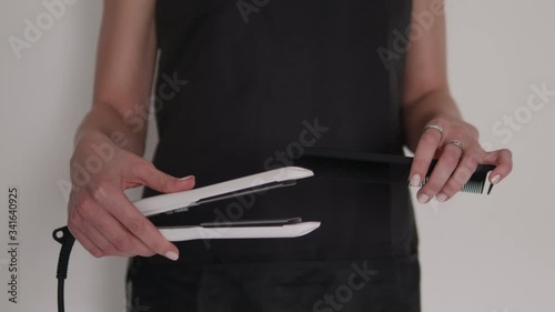 Detail shot of hairdresser woman's hands holding and demonstrating professional barber tools curling iron and comb with black apron background on hairstyling salon 