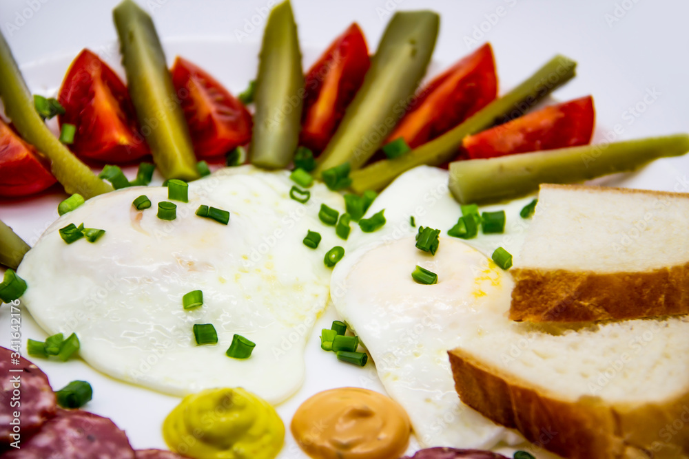 Delicious breakfast with eggs, salami, cucumbers, cherry tomatoes, slices of loaf, sauces and green onion on top on a white plate. Healthy and tasty meal for breakfast or lunch. Delicious breakfast with eggs, salami, cucumbers, cherry tomatoes, slices of loaf, sauces and green onion on top on a white plate. Healthy and tasty meal for breakfast or lunch.