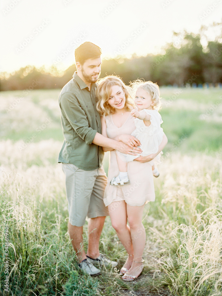 Fototapeta premium Portrait of a happy family father, mother, little daughter having fun together, father holding little daughter. Happy loving family walking outdoor in the light of sunset.