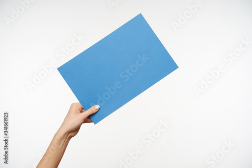 Indoor shot of young female's elegant hand being raised while keeping piece of light blue paper in it, passing it to someone while posing over white background