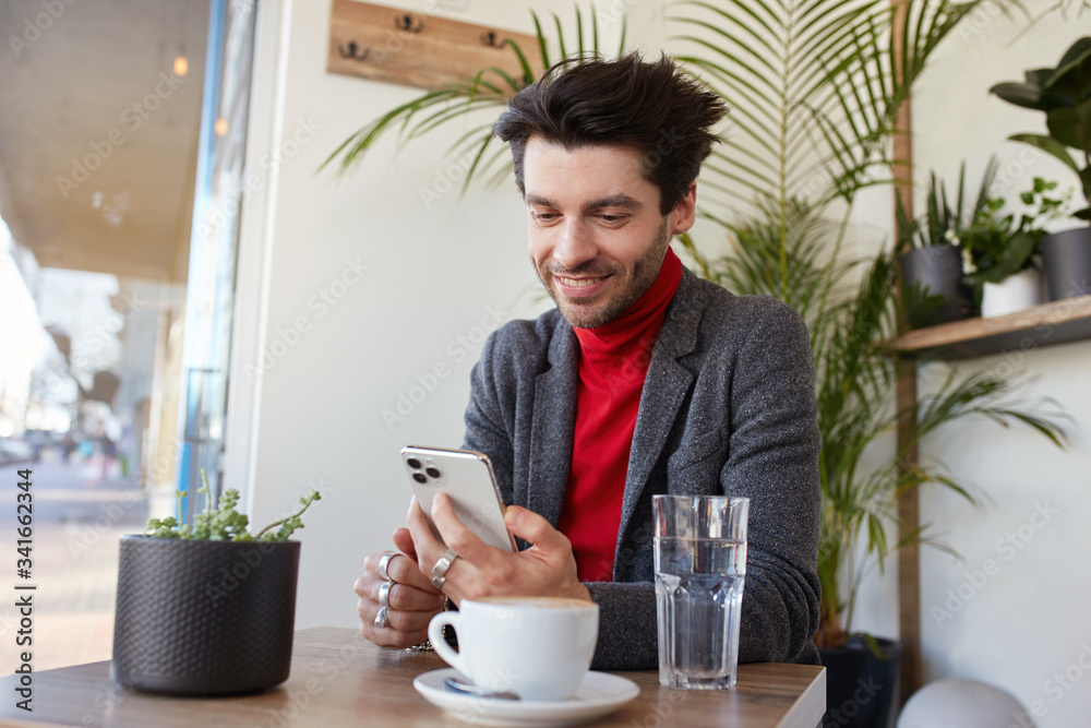 Cheerful young attractive brown haired unshaved male smiling pleasantly while looking on screen of his phone, having lunch break and sitting in city cafe Cheerful young attractive brown haired unshaved male smiling pleasantly while looking on screen of his phone, having lunch break and sitting in city cafe