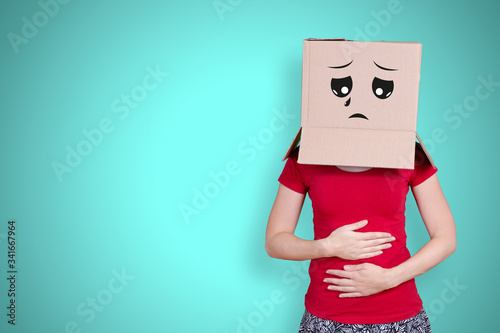 Person with cardboard box on its head and a sad face expression holding ist belly on turquoise background