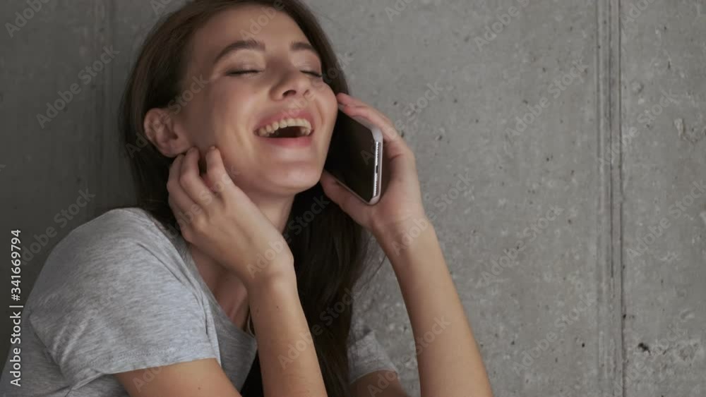 Joyful pretty brunette woman talking by smartphone and enjoying this moment while sitting near the wall at home Joyful pretty brunette woman talking by smartphone and enjoying this moment while sitting near the wall at home
