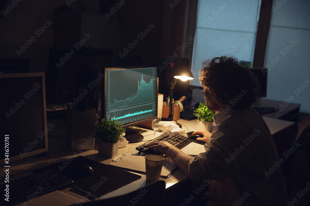 Working with graphs. Man working in office alone during COVID-19 quarantine, staying to late night. Young businessman, manager doing tasks with smartphone, laptop, tablet in empty workspace. Working with graphs. Man working in office alone during COVID-19 quarantine, staying to late night. Young businessman, manager doing tasks with smartphone, laptop, tablet in empty workspace.