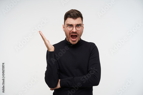 Bewildered young pretty short haired unshaved man raising emotionally hand while looking confusedly at camera with opened mouth, standing against white background