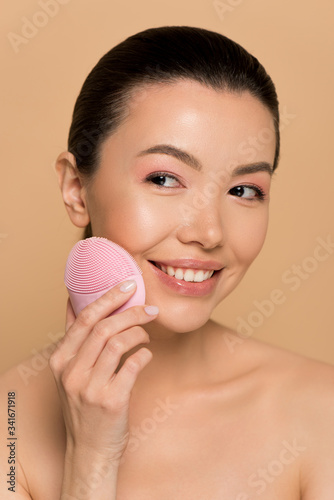 attractive nude asian girl using pink silicone cleansing facial brush isolated on beige