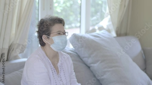 Happy beautiful older middle aged woman wearing protective mask relaxing sitting on sofa at home, stay at home health care COVID-19 pandemic situation.