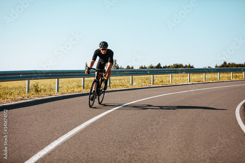 Active mature man in sport outfit riding bicycle on paved road in countryside. Experienced cyclist in black helmet and mirrored glasses regularly training outdoors.