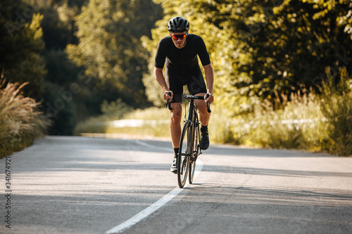 Fototapeta Naklejka Na Ścianę i Meble -  Strong sportsman with athletic body wearing black sport clothing, helmet and sunglasses practicing in cycling on fresh air. Concept of healthy hobby and regular trainings