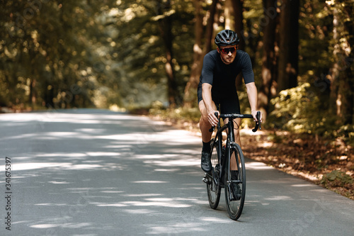 Professional cyclist in sport clothing and black helmet riding bike on paved road among summer forest. Bearded athlete in protective eyeglasses training outdoors.