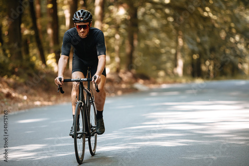 Mature man in sport clothing, protective helmet and sunglasses riding black professional bike with blur background of nature. Male cyclist enjoying summer days during sport activity. © Tymoshchuk