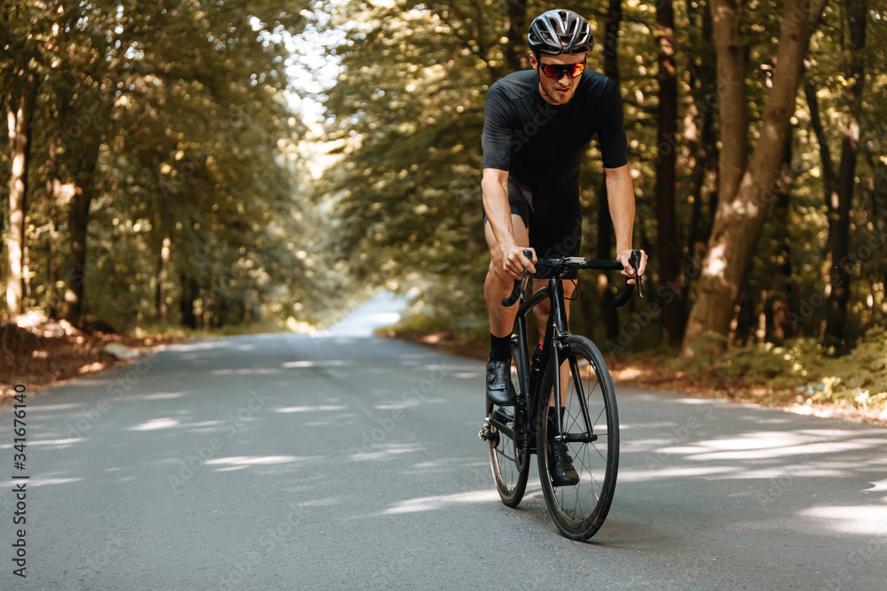 Fototapeta premium Tired strong athlete in black sport clothing training on bike during summer days. Mature man in helmet and eyewear actively riding bike outdoors.