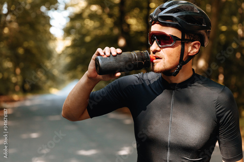 Fototapeta Naklejka Na Ścianę i Meble -  Portrait of exhausted sportsman in black helmet and mirrored glasses drinking cold water from sport bottle while standing in green forest. Mature man having break during long ride.