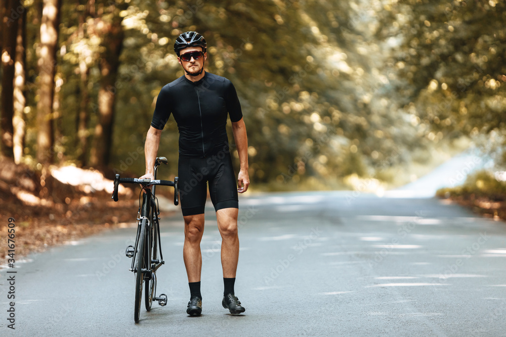 Strong male cyclist in sportswear and protective helmet walking with his bike at paved road to take a break after riding. Concept of active and healthy lifestyle