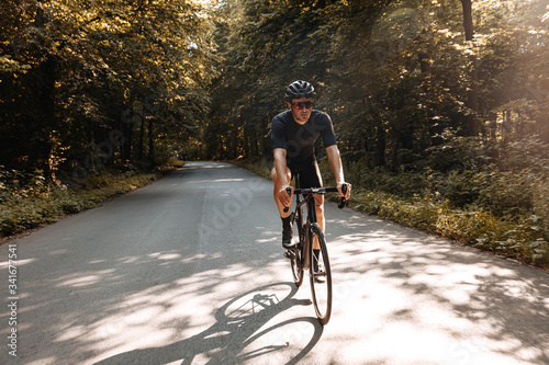 Fototapeta Naklejka Na Ścianę i Meble -  Active bearded athlete in sport clothing riding bike on paved road during sunny days. Strong sportsman in protective helmet and glasses training regularly outdoors.